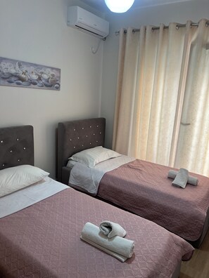 A hotel room with two beds, a wall-mounted air conditioner, a painting, and a window with curtains. - Cozy 1-bedroom apartment with sea view (Saranda)