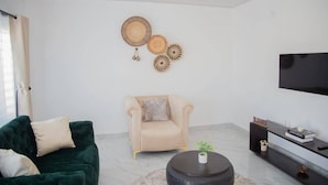 A living room with a dark green sofa, a beige armchair, a round coffee table, and a flat-screen TV mounted on the wall.