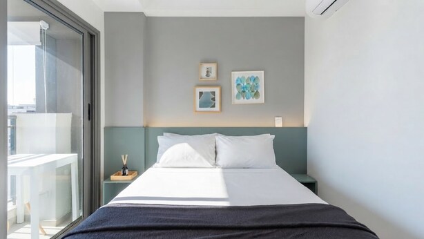 A hotel room with a bed, a nightstand, a small table with a lamp, and a window with a view of the outside. - Studio com Piscina perto do metrô Vila Mariana (São Paulo)