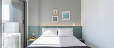 Quarto de hotel com cama, mesa de cabeceira, pequena mesa com abajur e janela com vista para o exterior.