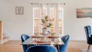 A dining area with a glass table, blue chairs, a vase with flowers, and a painting on the wall.