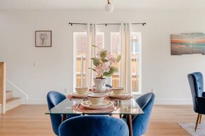 A dining area with a glass table, blue chairs, a vase with flowers, and a painting on the wall.
