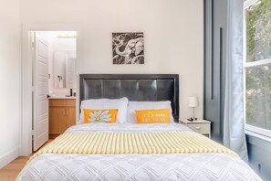 Une chambre à coucher comprenant un lit, une table de chevet, une fenêtre avec des rideaux, une porte et un tableau accroché au mur.