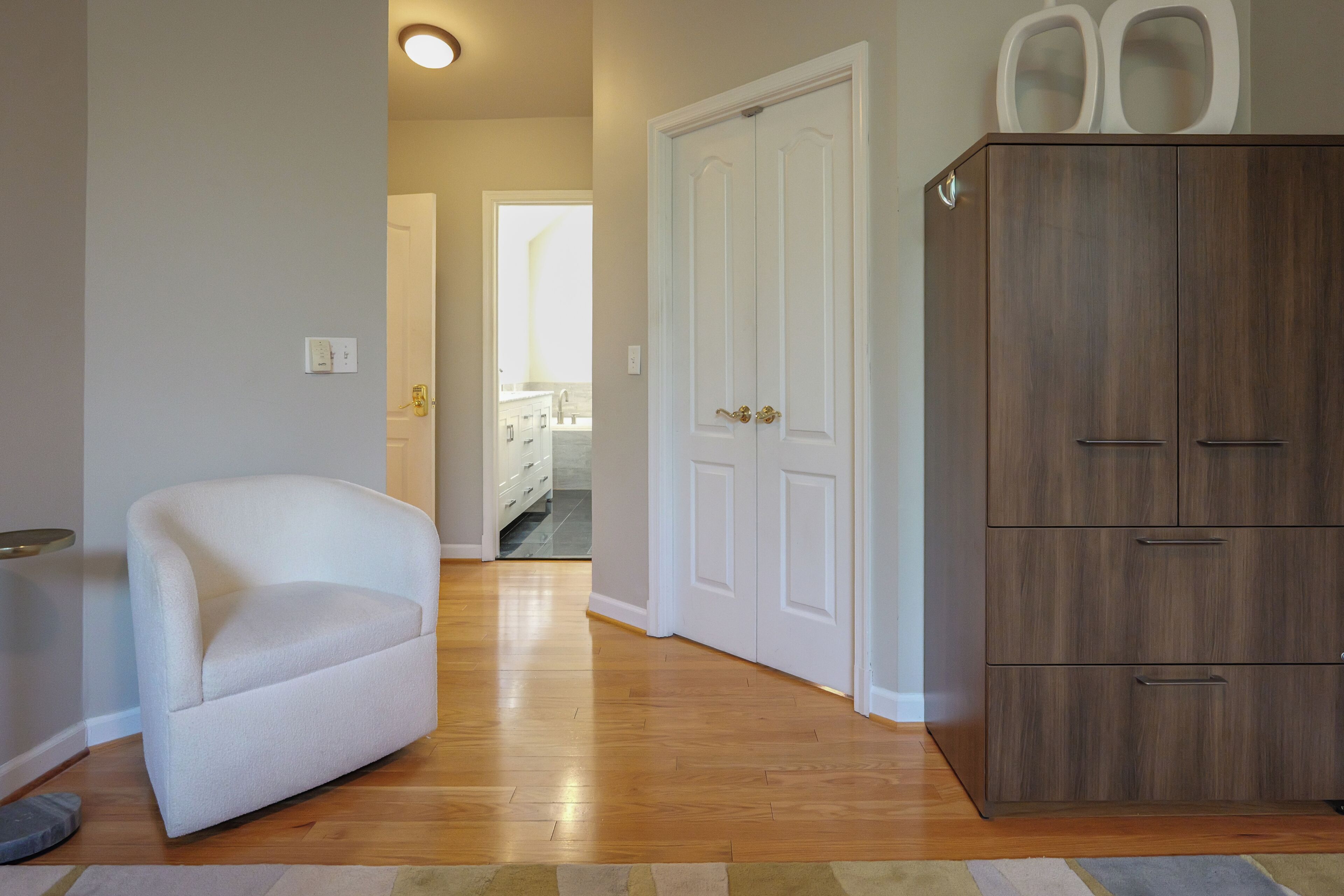 A room with a white armchair, wooden flooring, a wardrobe, and a door leading to a bathroom.