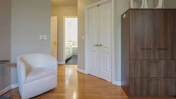 A room with a white armchair, wooden flooring, a wardrobe, and a door leading to a bathroom.