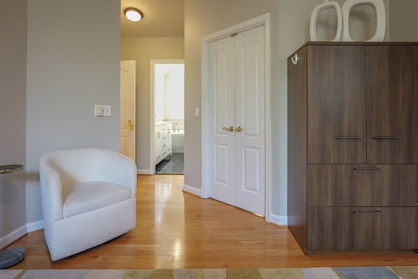 A room with a white armchair, wooden flooring, a wardrobe, and a door leading to a bathroom.