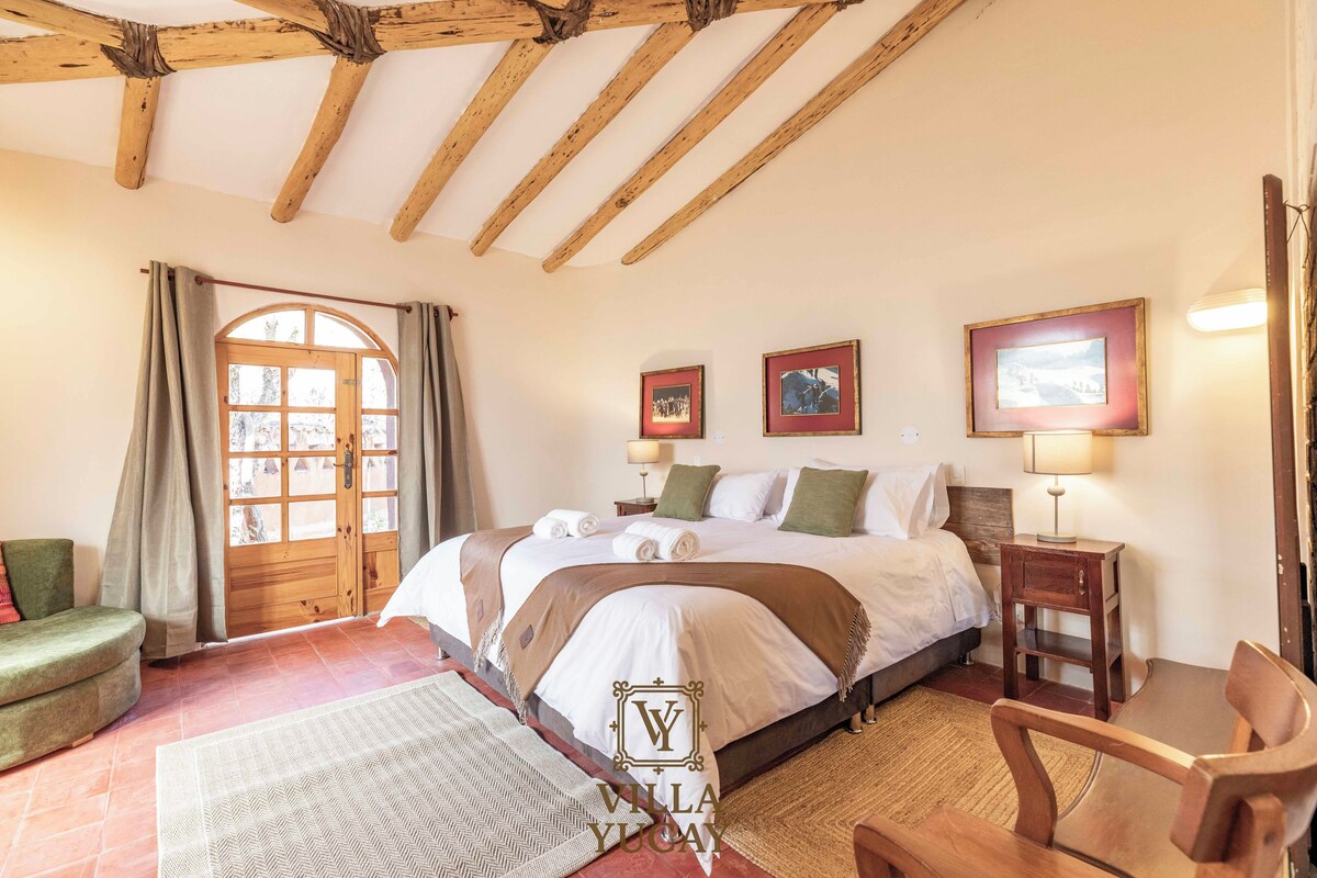 Habitación de hotel con techo de madera, cama con ropa blanca, mesita de noche de madera, una silla y un ventanal grande con cortinas.