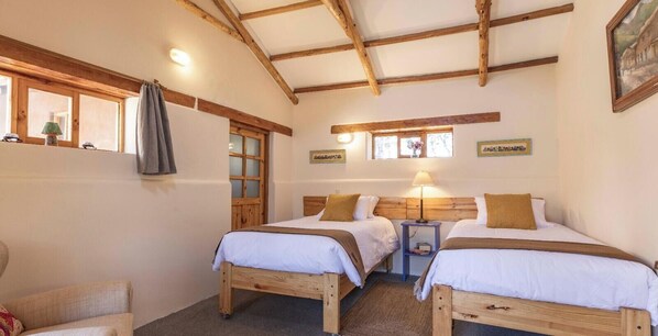 A room with two wooden beds, a small table, a chair, and a window with curtains.