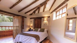 Un dormitorio con techo de madera, una cama con ropa blanca, una mesita de noche con lámpara y una ventana con vistas a la vegetación.