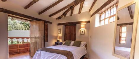 A bedroom with a wooden ceiling, a bed with white bedding, a nightstand with a lamp, and a window with a view of greenery.