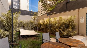 An outdoor seating area with a table and chairs, surrounded by dense foliage and a building wall. - Studio com Piscina Perto do Metr Vila Mariana (São Paulo)
