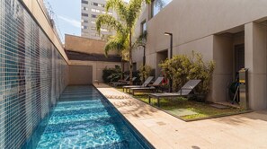 A swimming pool with a tiled edge, surrounded by lounge chairs and greenery, with a building in the background. - Studio com Piscina Perto do Metr Vila Mariana (São Paulo)