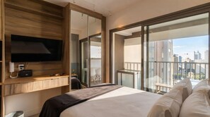 A modern hotel room with a large bed, a flat-screen TV mounted on a wooden panel, a balcony with a city view, and a sliding glass door. - Studio com Piscina Perto do Metr Vila Mariana (São Paulo)