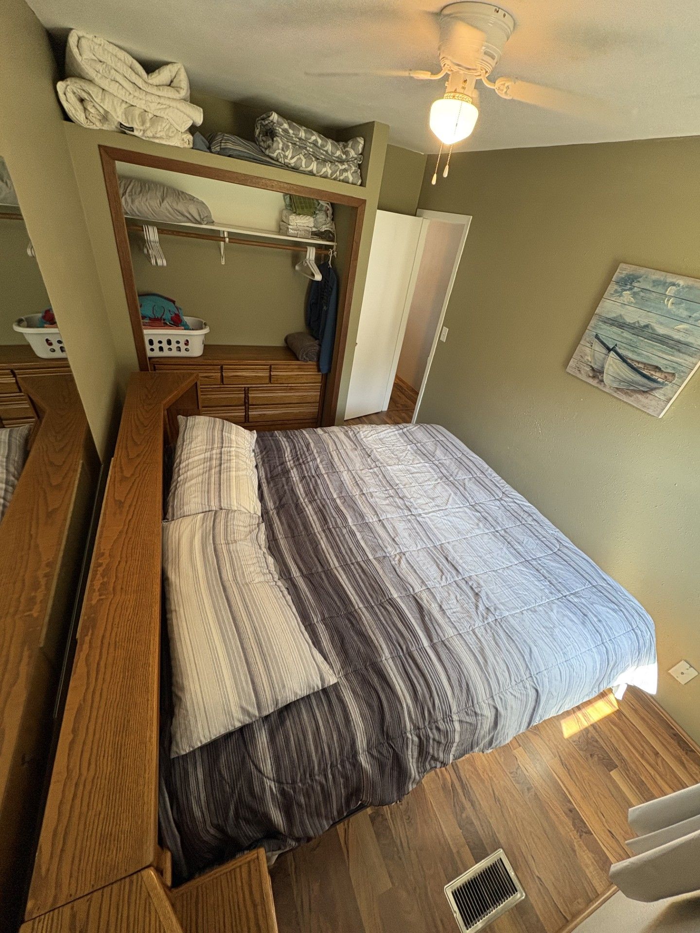 A bedroom with a bed, wooden headboard, a ceiling fan, a wall-mounted painting, and a storage unit above the bed.