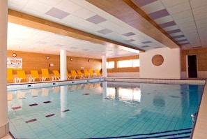 Ein Hallenbad mit gefliestem Boden und Wänden, Holzbalken und gelben Stühlen am Rand.