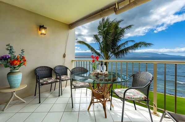 A balcony with a glass table, wicker chairs, a vase with flowers, and a view of the sea.