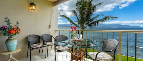 A balcony with a glass table, wicker chairs, a vase with flowers, and a view of the sea.