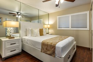 A bedroom with a bed, bedside tables, a mirror, a ceiling fan, and a window with blinds. - Bright 4BR, 2 Condos for 12 at Maalaea Kai! (Wailuku)
