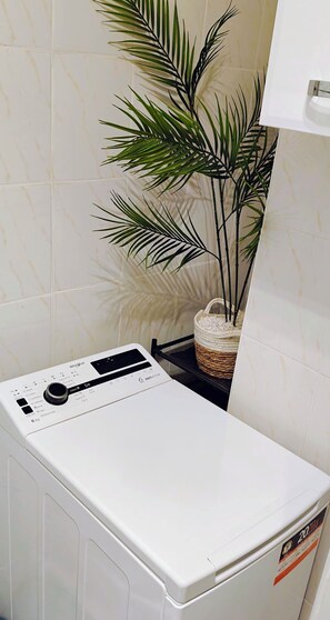 A front-loading washing machine with a control panel, a potted plant, and a basket of white items on a shelf. - Blue Dream - Warsaw Ochota Apartment - Easy Access to Chopin Airport (Warszawa)