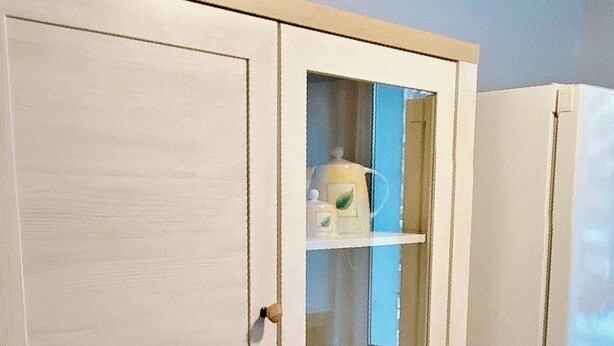 Valkoinen kaappi, jossa on useita laatikoita ja hyllyjä, lasiovilla varustettu osa, jossa on pullo ja lasi, sekä valkoinen jääkaappi.