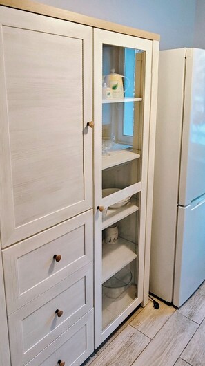 A white cabinet with multiple drawers and shelves, a glass-fronted section with a bottle and glass, and a white refrigerator. - Blue Dream - Warsaw Ochota Apartment - Easy Access to Chopin Airport (Warszawa)