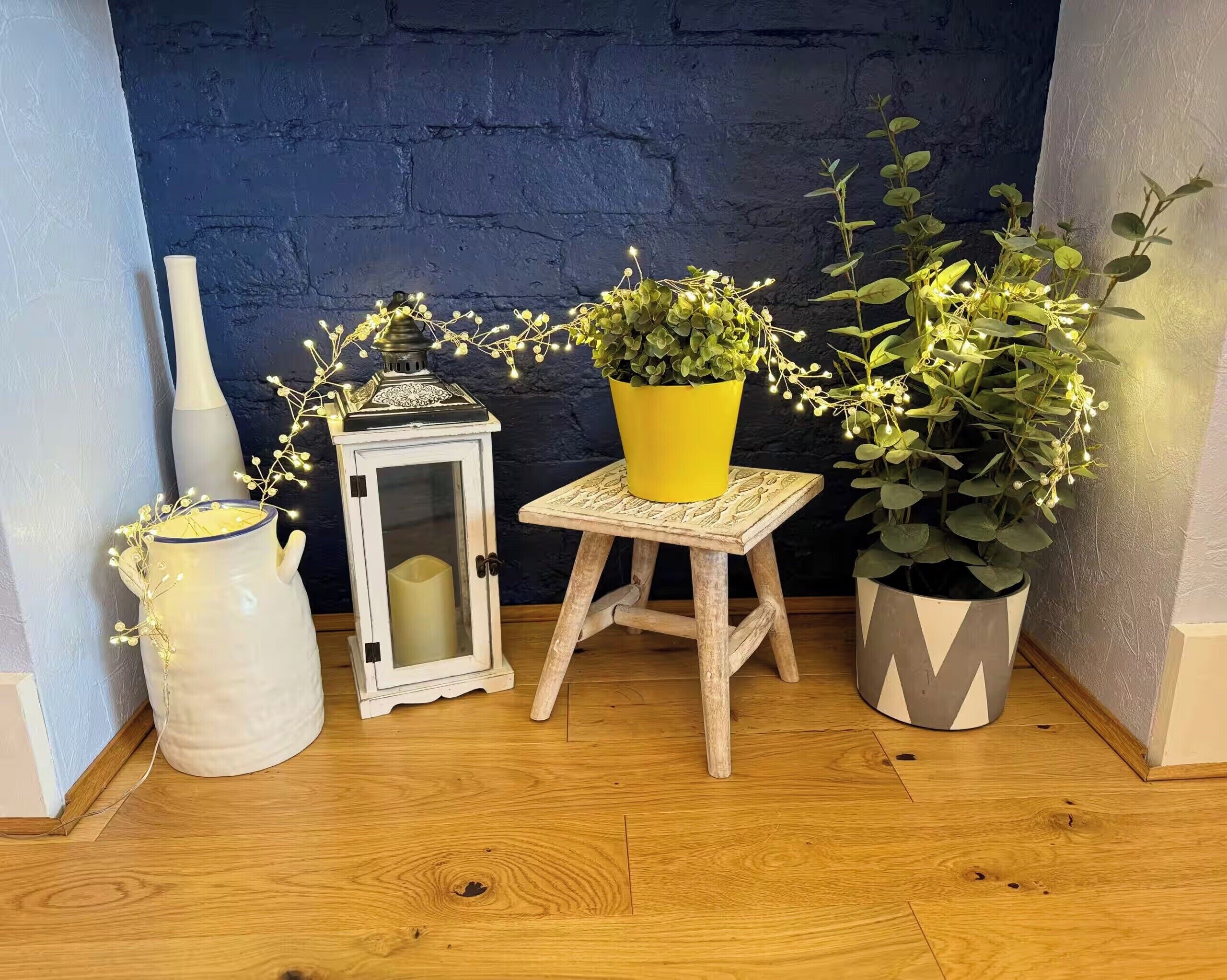 A corner with a blue wall, a white vase, a lantern, a small wooden stool, and two potted plants.