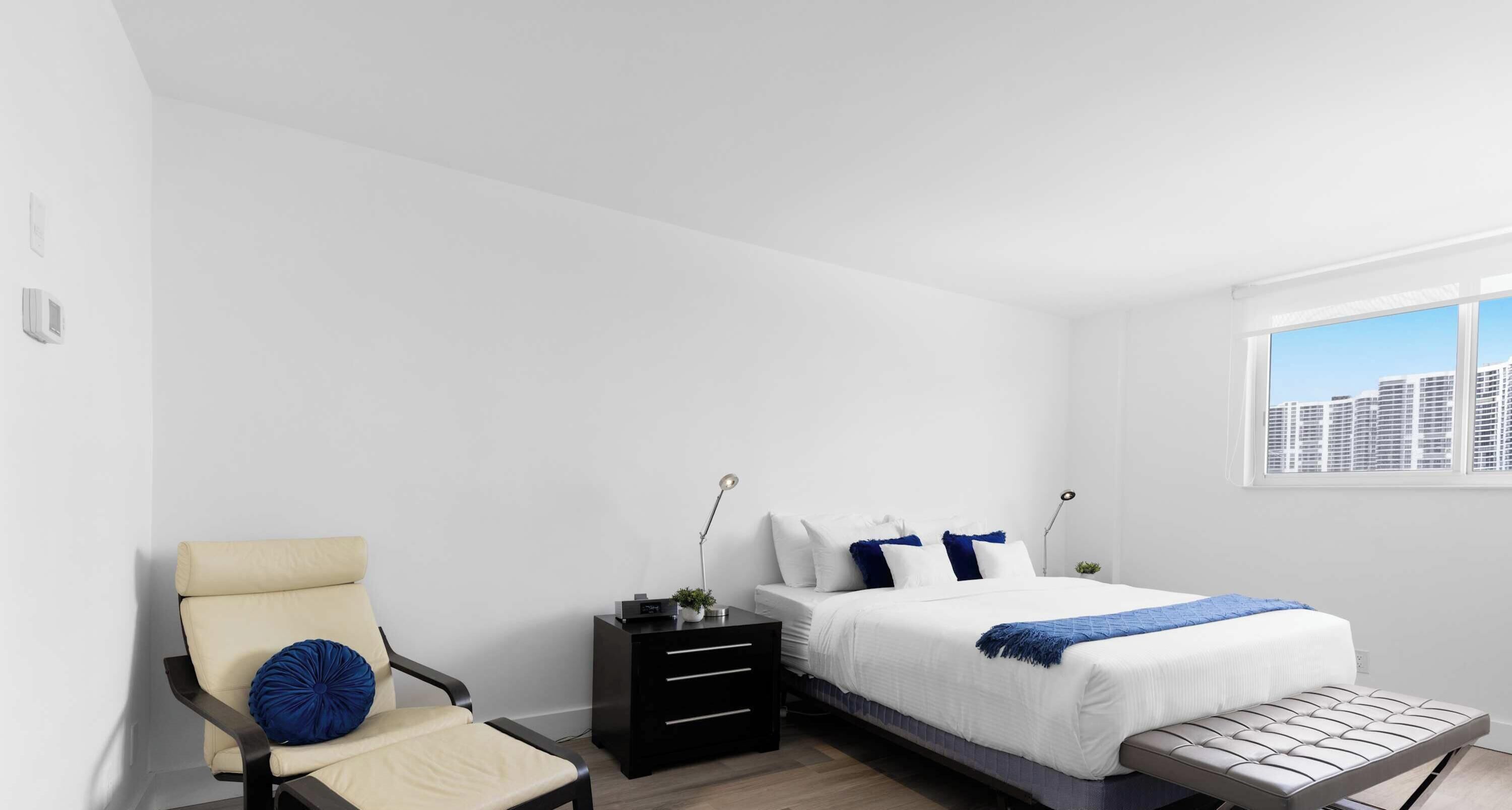 Un dormitorio moderno con cama, mesita de noche, silla y vista a la ciudad desde una ventana.