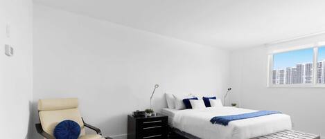 Un dormitorio moderno con cama, mesita de noche, silla y vista a la ciudad desde una ventana.