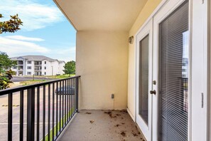 Un balcon avec une balustrade noire, une porte avec une fenêtre, et une vue sur un parking et des bâtiments en arrière-plan.