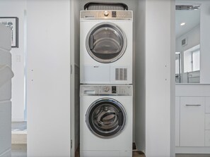 A stacked washer and dryer unit in a modern, minimalistic laundry room with a clean, white interior. - Sandy Strides - Whangamata Holiday Home (Whangamata)