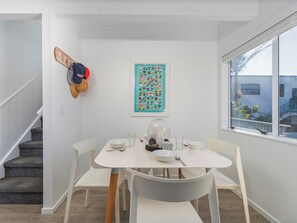 A dining area with a table set for two, a staircase, a wall-mounted hat rack, and a window with a view of a building. - Sandy Strides - Whangamata Holiday Home (Whangamata)