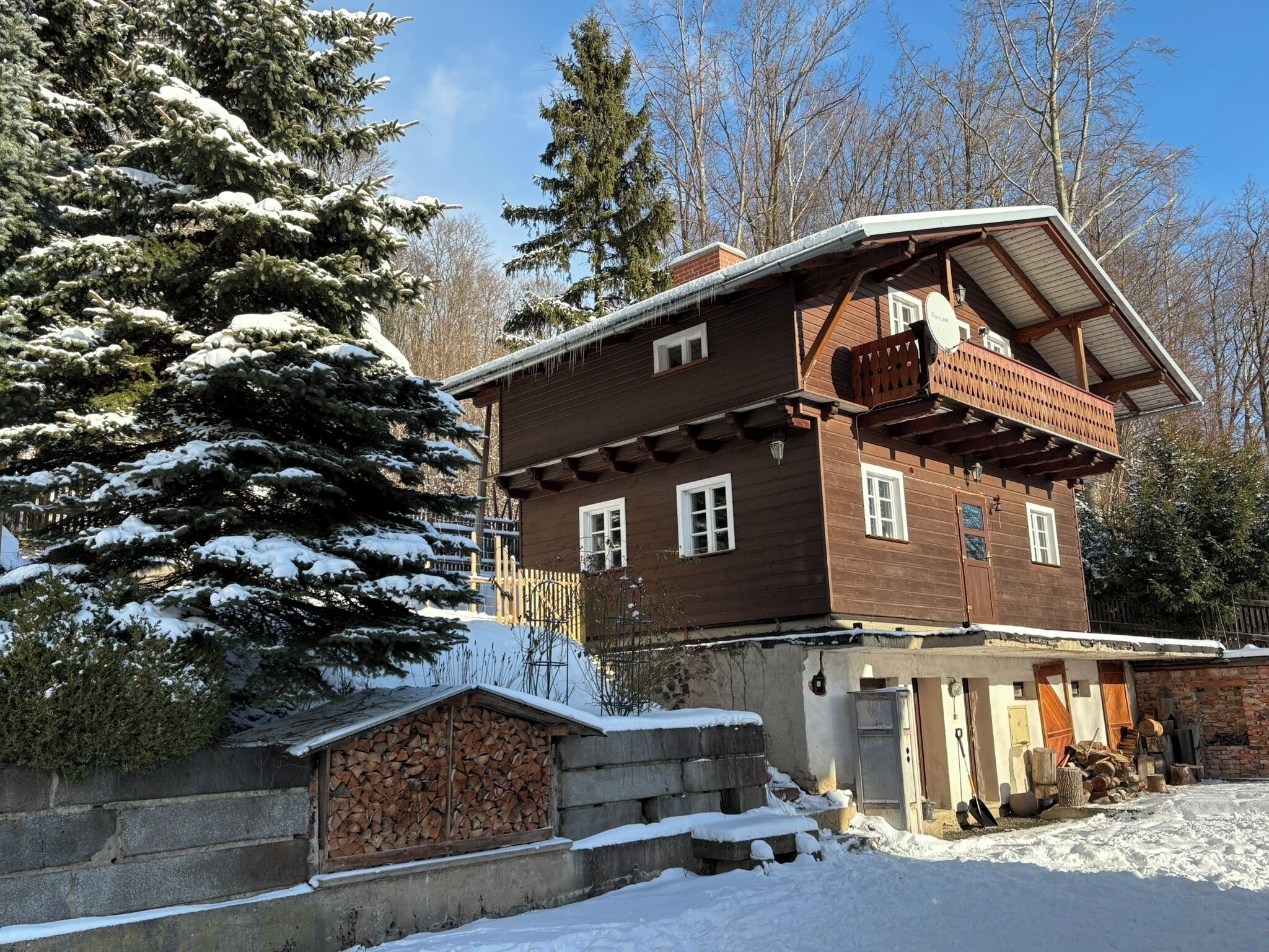 Une maison en bois à deux étages, dotée d’un balcon, entourée d’arbres couverts de neige et sous un ciel dégagé.
