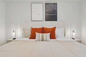 A neatly made bed with white bedding and two orange pillows, flanked by two bedside lamps.