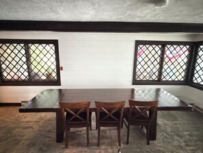 A dining area with a long table and three chairs, a white wall, and a window with a lattice design.