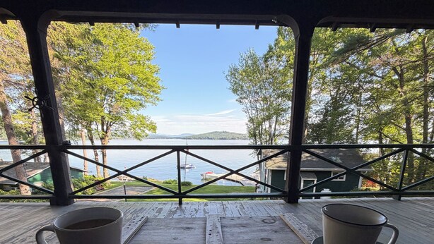 En træterrasse med to kopper, et træbord og udsigt over en sø med både og træer.