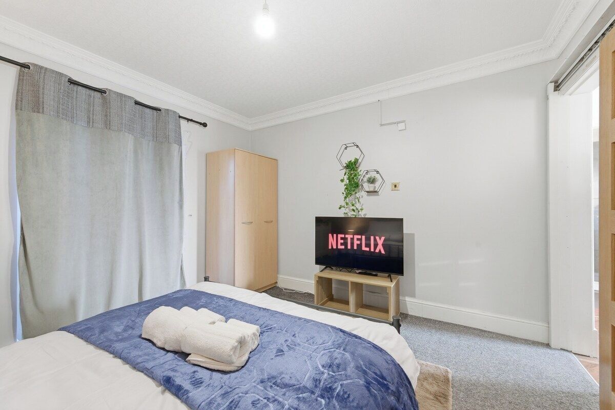 Ein Hotelzimmer mit Bett, einem Fernseher, der 'NETFLIX' zeigt, einem Kleiderschrank und einer Pflanze an der Wand.