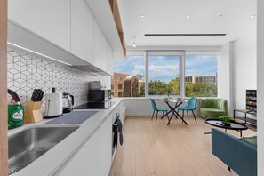 A modern kitchen with a large window, a dining area with a table and chairs, and a view of buildings outside. - Brentford 1 BR Corporate Relocation | Free Parking | Workspace | Gym | AC (Brentford)