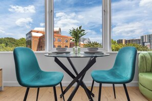 A dining area with a glass table, two teal chairs, and a view of a building through large windows. - Brentford 1 BR Corporate Relocation | Free Parking | Workspace | Gym | AC (Brentford)