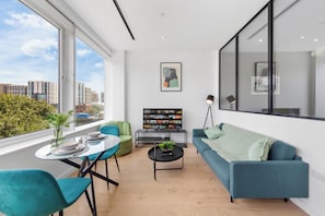A modern living room with a large window, a sofa, a dining table, and a TV stand with a movie poster. - Brentford 1 BR Corporate Relocation | Free Parking | Workspace | Gym | AC (Brentford)