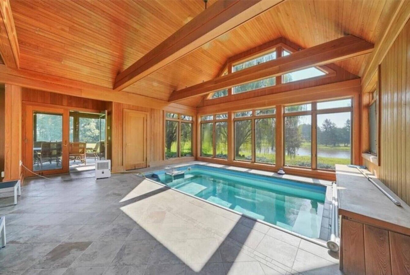 A spacious indoor swimming pool with a view of the outdoors, wooden ceiling beams, and a large window.