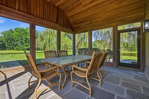 A covered patio with a table and chairs, large windows, and a view of a green landscape. - Spacious ADA 6BR Home for Groups w. Private Pond (Goshen)