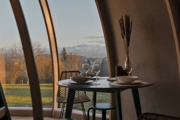 A dining area with a table set for two, a view of a mountainous landscape through large windows, and a tall vase with reeds.
