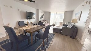 Quarto de hotel moderno com área de refeições, sofá, televisão e uma grande janela.