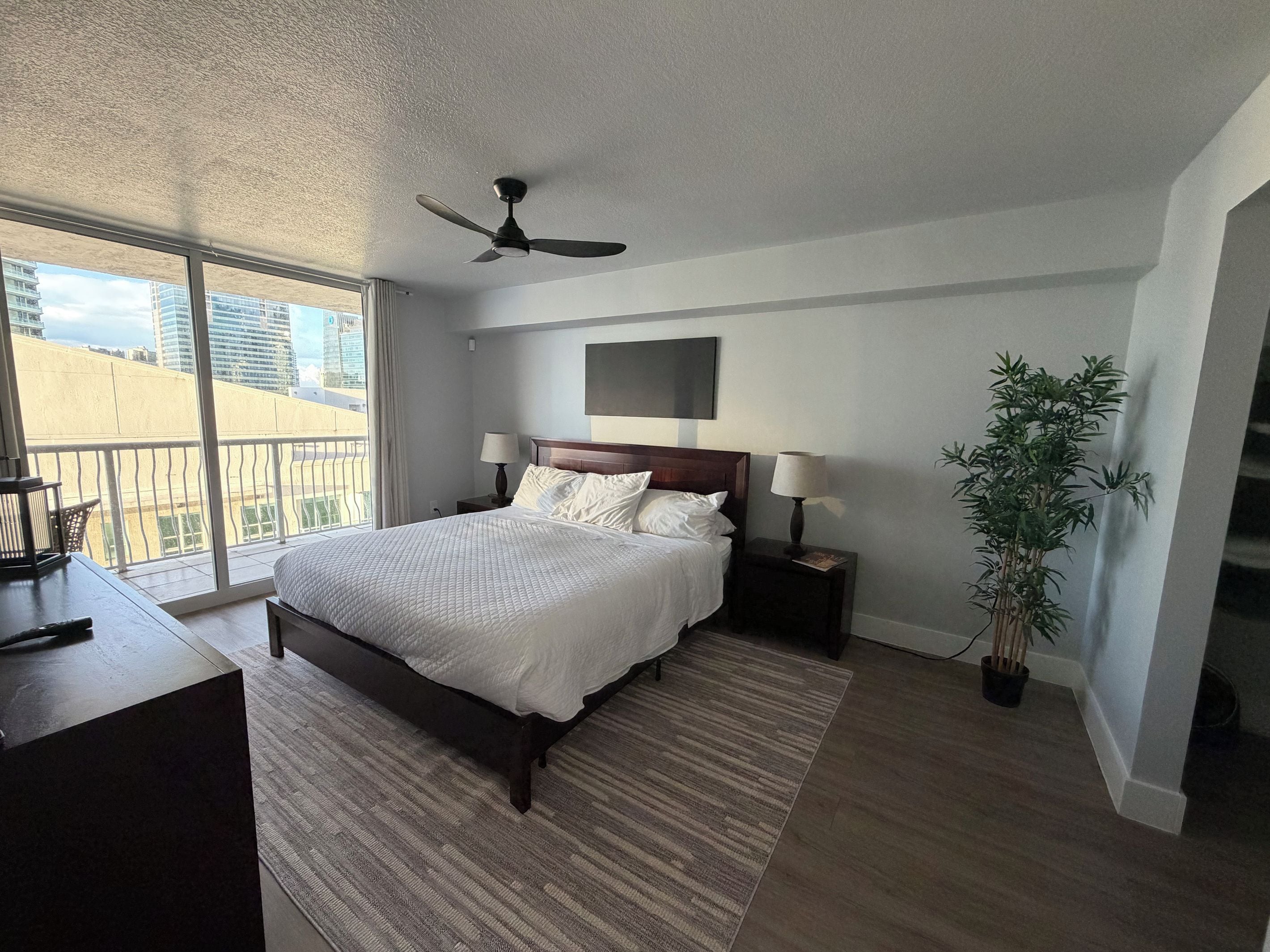 A bedroom with a large bed, bedside tables, a ceiling fan, a potted plant, and a view of the city.