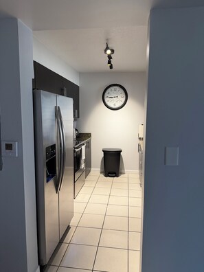 A modern kitchen with a stainless steel refrigerator, oven, and microwave. A clock is mounted on the wall, and a trash bin is visible. - Haven on Brickell Bay (Miami)