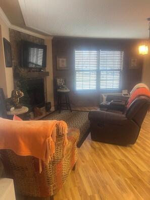 Ein Wohnzimmer mit Kamin, einem darüber montierten Fernseher, einer Couch, einem Liegestuhl und einem gemusterten Teppich.