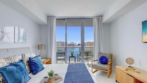 A modern bedroom with a large bed, a chair, a bedside table, a floor lamp, and a view of a cityscape through a balcony. - 2BR - Bay View Fitness Center & Private Balcony (Hallandale Beach)