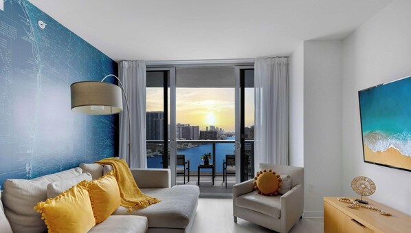 A modern living room with a sofa, armchairs, a dining table, and a view of a cityscape and water body. - 2BR - Bay View Fitness Center & Private Balcony (Hallandale Beach)