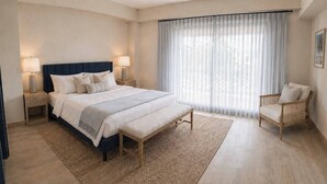 Quarto de hotel com uma cama grande, abajures, um banquinho, uma cadeira e uma janela com cortinas finas.