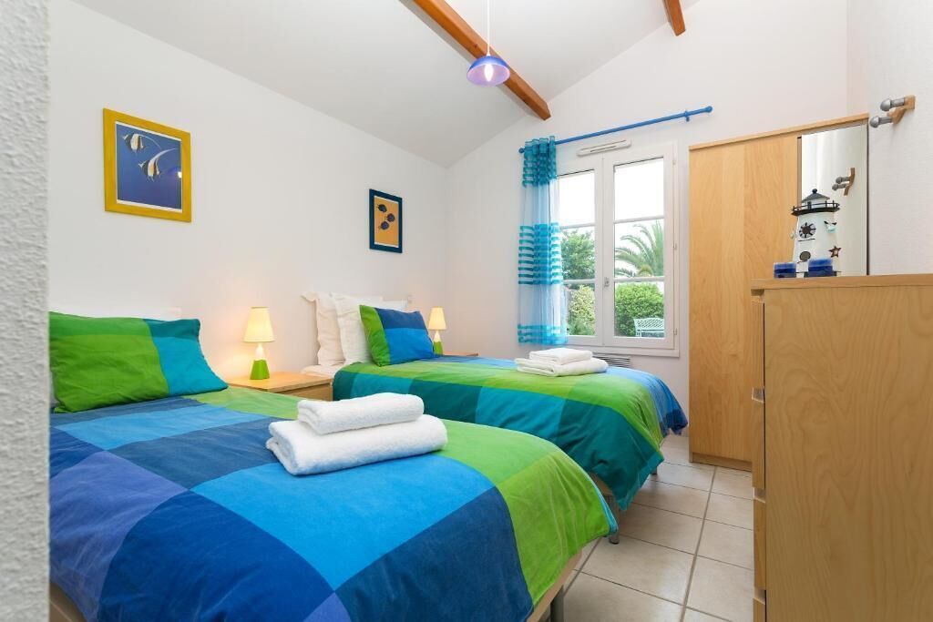 A bedroom with two beds, a wooden wardrobe, a window with curtains, and a small table with a lamp.